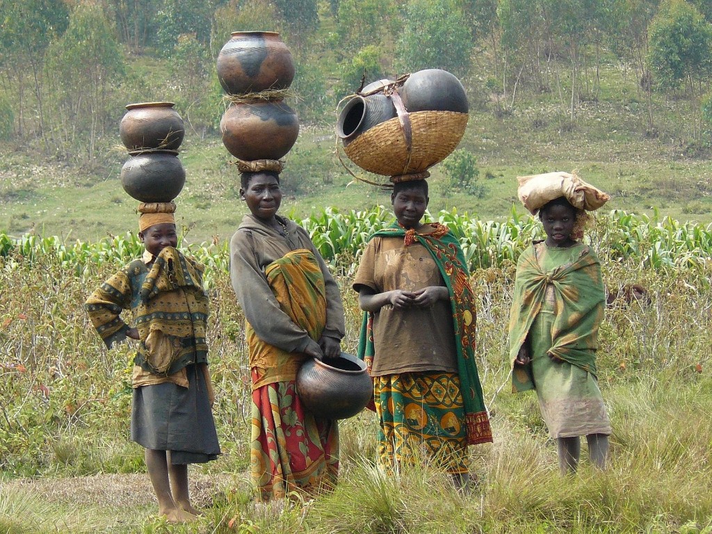 Batwa community members carrying traditional pottery in a rural Ugandan highland landscape